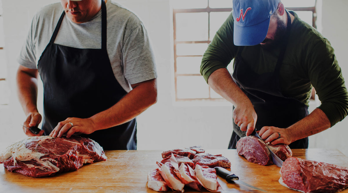 Two butchers cutting beef primals into steaks at a wooden butcher block in a well-lit facility
