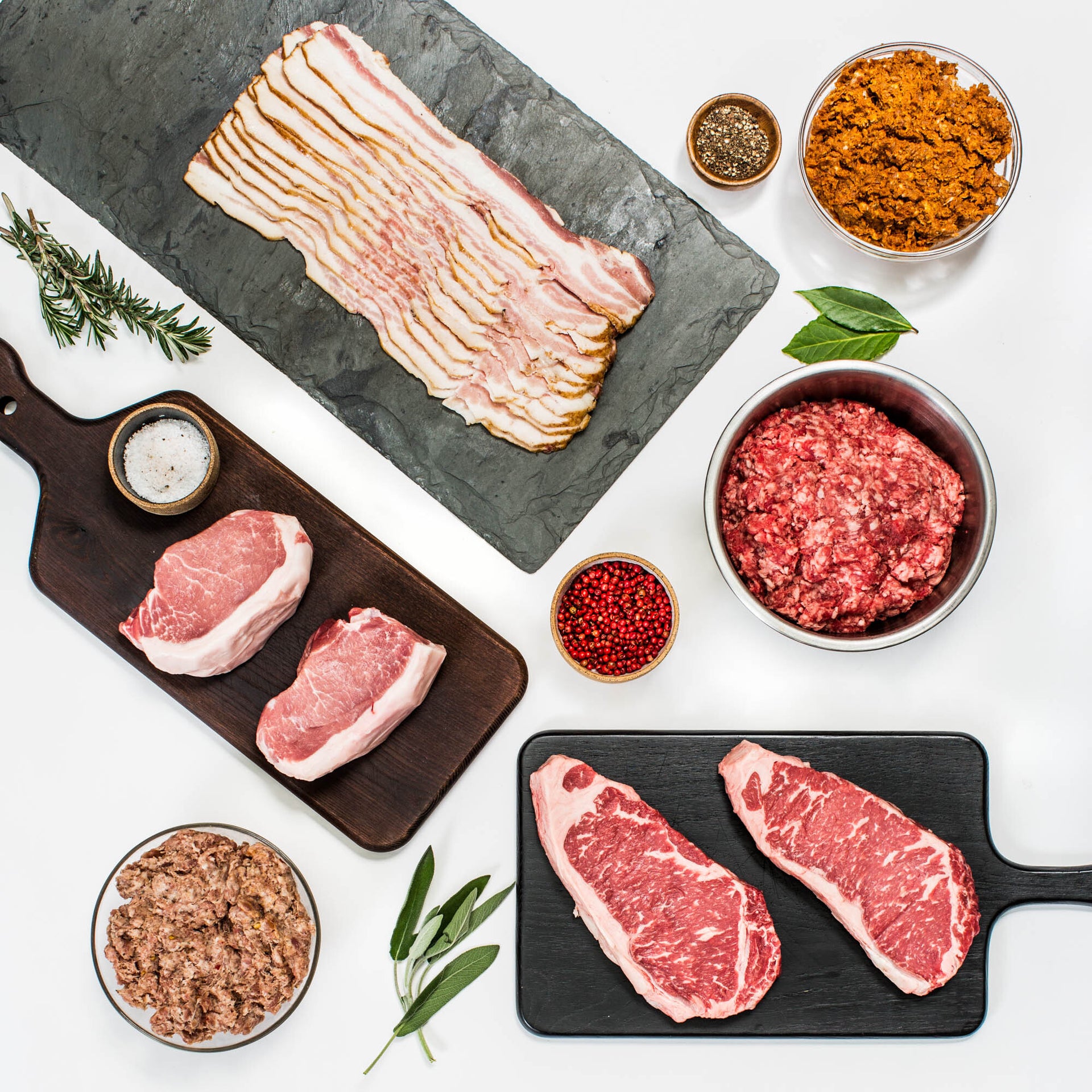 Assorted raw pasture-raised meats including steaks, bacon, pork loin, ground beef, and sausage with herbs and seasonings